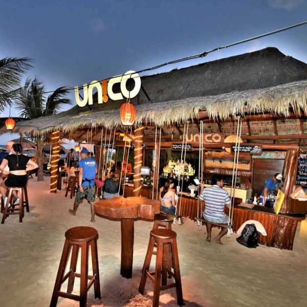people sit on stools around small tables set right on sand. A bar with a palapa style roof serves patrons seated on swings hung from the roof