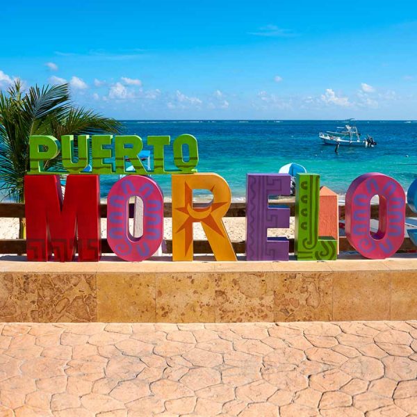 a colorful sign on the beach reads, Puerto Morelos. Boats float in the blue waters of the Caribbean beyond the beach